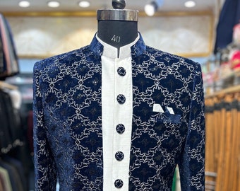 May include: A navy blue velvet jacket with a white, intricate, repeating geometric pattern and a mandarin collar. The jacket features a central white placket with seven matching navy blue buttons, long sleeves, and a small pocket square detail.
