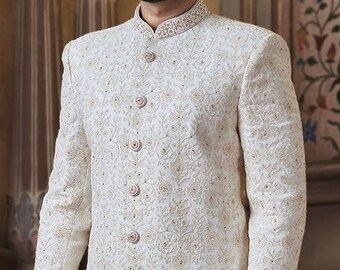 Puede incluir: Un sherwani blanco bordado para hombre con un turbante blanco. El sherwani tiene un cuello alto y un cierre de botones en la parte delantera. El bordado es intrincado y detallado.