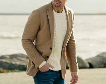 Op de afbeelding: Een man draagt ​​een bruine blazer over een witte kabelbreistruien en blauwe jeans. Hij staat buiten met een waterpartij op de achtergrond.