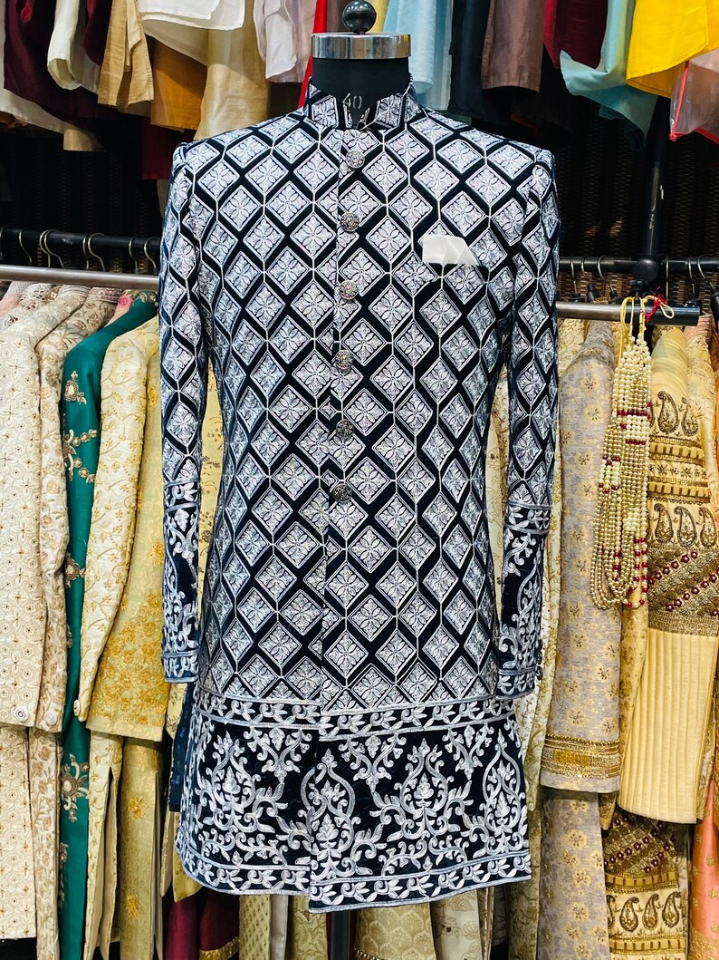May include: A black and white patterned jacket with a mandarin collar and silver buttons. The jacket features a geometric design and ornate embroidery along the hem and cuffs. A white pocket square is visible.
