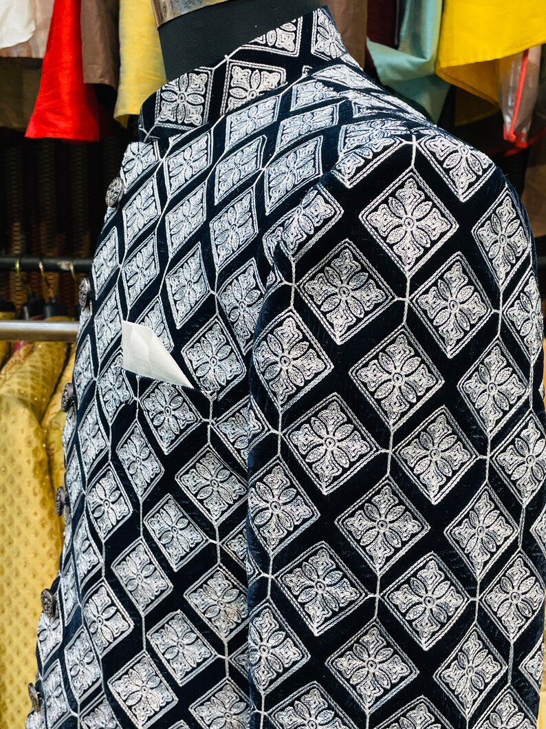 May include: A black velvet jacket with a diamond pattern embroidered in white. The jacket has a mandarin collar, decorative buttons, and a white pocket square. The pattern is a repeating geometric design with floral elements.