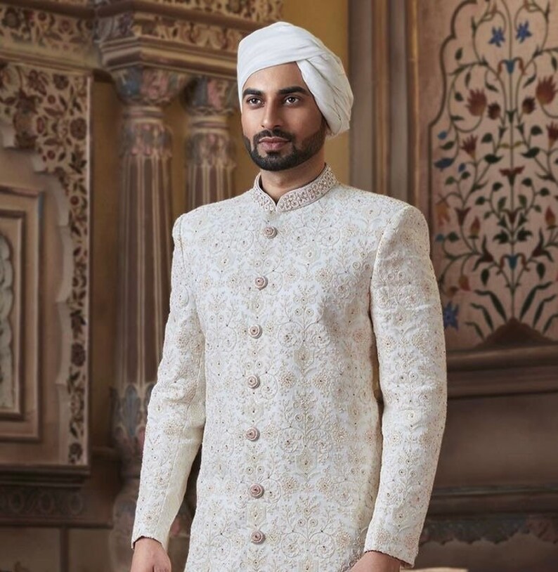 May include: A white embroidered men's sherwani with a white turban. The sherwani has a high collar and a button-up front. The embroidery is intricate and detailed.
