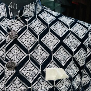 May include: Close-up of a black jacket with a geometric diamond pattern embroidered in white. The jacket features ornate silver buttons and a white pocket square. The jacket is displayed on a mannequin.