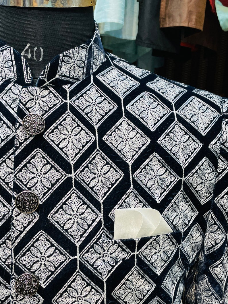 May include: Close-up of a black jacket with a geometric diamond pattern embroidered in white. The jacket features ornate silver buttons and a white pocket square. The jacket is displayed on a mannequin.