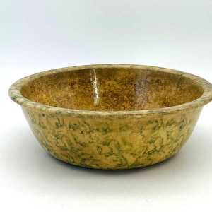 May include: A round, speckled bowl with a wide rim. The bowl has a mottled pattern of green and brown hues, with a light-colored interior. The bowl is made of ceramic and is suitable for serving food.