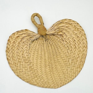 May include: A heart-shaped hand fan made of woven natural fibers. The fan has a loop handle and a tightly woven pattern. The color is a light tan, and the fan is set against a white background.