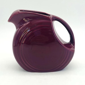 May include: A deep burgundy ceramic pitcher with a rounded body and a large handle. The pitcher has a spout for pouring and a glossy finish. The design features concentric rings around the base.