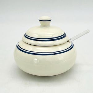 May include: A cream-colored ceramic sugar bowl with a rounded body and a matching lid. The bowl and lid are decorated with two thin, dark blue horizontal stripes. A clear, transparent acrylic spoon is inserted into the side of the bowl.