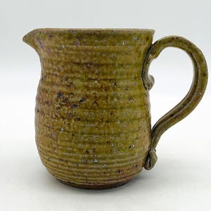 May include: A ceramic pitcher with a textured, ribbed exterior surface in shades of olive green and brown. The vessel features a small pouring spout, a curved handle attached to the side, and a speckled finish throughout the clay body.