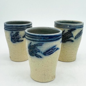 May include: Three ceramic tumblers with a speckled beige finish. Each tumbler features a dark blue rim and a stylized blue bird design. The tumblers are of similar size and shape, suitable for beverages.