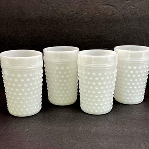 May include: A set of four white milk glass tumblers featuring a raised hobnail texture across the exterior surface. Each cylindrical glass has a smooth rim and a consistent opaque finish, arranged in a group of four identical drinking vessels.