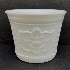 May include: A white milk glass planter featuring a vertical ribbed pattern around the exterior, a decorative embossed cartouche design on the front, and a scalloped rim detail. The vessel has a wide opening and a tapered cylindrical shape.