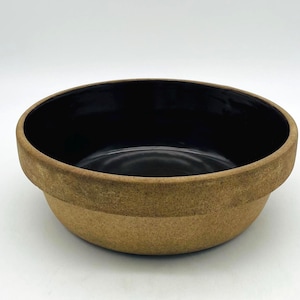 Könnte beinhalten: Eine runde, beige Keramikschüssel mit dunklem Innenraum. Die Schüssel hat einen breiten Rand und eine leicht strukturierte Oberfläche. Der Innenraum ist glänzend schwarz und kontrastiert mit der matten Außenseite. Die Schüssel wird wahrscheinlich zum Servieren oder Mischen verwendet.