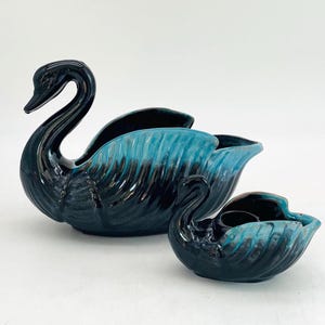 May include: Two ceramic swan figurines. The larger swan is dark blue with teal accents, and the smaller swan has a similar color scheme. Both have a glossy finish and are positioned on a white surface. The swans are decorative items.