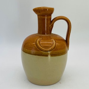 Może przedstawiać: Dwukolorowy ceramiczny dzban z uchwytem. Górna część jest w ciepłym brązie, a dolna w jasnobeżowym kolorze. Dzban ma dzióbek i zakrzywiony uchwyt. Na ramieniu dzbana znajdują się elementy dekoracyjne.