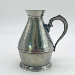 May include: A silver-toned metal pitcher with a wide, rounded base that tapers toward a flared rim. The vessel features a single curved handle attached to the side, a smooth polished finish, and a raised circular base ring for stability.
