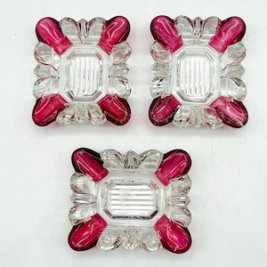 May include: Three clear glass decorative squares with a rectangular center and a scalloped edge. Each square has a vibrant magenta accent on each corner. The squares are arranged on a white background.