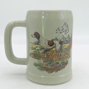 May include: A light beige ceramic beer mug featuring a detailed illustration of a white and brown spotted hunting dog standing in a field of tall grass with trees in the background. The mug has a sturdy handle and a smooth, cylindrical body.