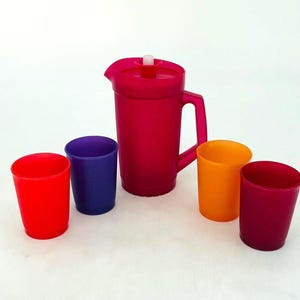 Könnte beinhalten: Ein leuchtend pinkfarbener Kunststoffkrug mit weißem Deckel und Griff, zusammen mit vier bunten Bechern. Die Becher sind rot, lila, orange und dunkelrosa. Ein Getränkeset.