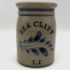May include: A cylindrical stoneware crock with a speckled tan finish and a dark blue painted leaf design. The text "SEA CLIFF" is embossed in an arc above the leaf, and the letters "L.I." are embossed in a straight line near the base.
