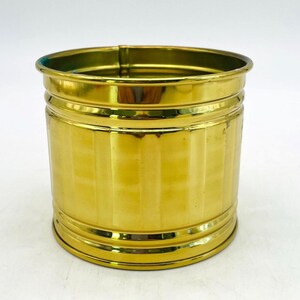 May include: A cylindrical, gold-colored metal container with vertical ribbed detailing. The container has a rounded top and base, and it is set against a white background. The container is likely a decorative item or a small planter.