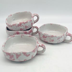 Peut inclure: Trois bols en céramique avec anses, présentant une base blanche avec un motif tacheté rose. Les bols ont une forme organique irrégulière avec une finition lisse et brillante. Deux bols sont empilés à gauche, un autre est posé à droite.