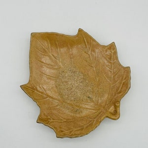 May include: A ceramic leaf-shaped dish in a light brown color. The dish has a textured surface that resembles the veins of a leaf.