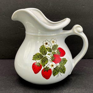 May include: White ceramic pitcher with a curved spout and a rounded body. The pitcher features a strawberry design with red berries, green leaves, and white flowers. The handle is a curved, elegant design.