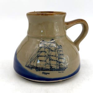 Könnte beinhalten: Ein beigefarbener Keramikbecher mit breitem, abgerundetem Boden und einem Henkel. Der Becher zeigt eine blaue Strichzeichnung eines Segelschiffs mit dem darunter gedruckten Schriftzug "Clipper" in Blau. Der untere Teil ist dunkelblau glasiert.