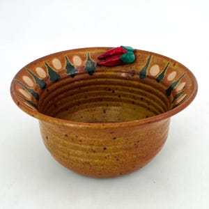 May include: A handmade ceramic bowl with a speckled brown exterior and a decorative rim. The rim features a pattern of alternating light and dark shapes, with a cluster of red chili peppers and green leaves. The bowl is ideal for serving.