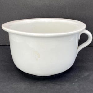 May include: A white ceramic chamber pot featuring a smooth, rounded basin with a wide, flat rim and a single curved handle on the side. The exterior surface is plain white with a subtle, light-colored blemish on the front of the bowl.