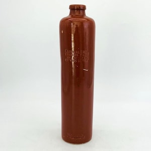 May include: A tall, cylindrical brown ceramic bottle with a smooth, glossy finish and a narrow neck. Embossed text on the front reads: FEDERAL LAW FORBIDS SALE OR RE-USE OF THIS BOTTLE. The bottle has a flat base and a small, rounded rim at the top.