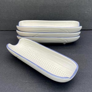 May include: A stack of four white ceramic corn-shaped dishes with a blue trim, and one dish in front. The dishes have a textured interior and are designed to hold corn on the cob. The background is a dark gray.