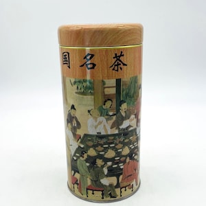 May include: A cylindrical tea tin with a wooden lid. The tin features a detailed illustration of a gathering of people around a table, with Chinese characters on the lid. The overall color scheme is muted with a focus on earth tones.