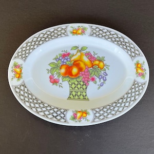 May include: Oval serving dish with a white center featuring a colorful fruit basket design. The rim has a gray and white lattice pattern with fruit accents. A decorative piece for serving or display.
