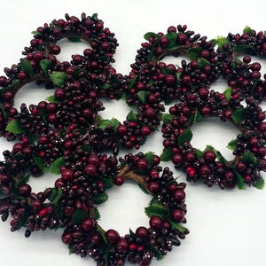 May include: A collection of small, round decorative wreaths. Each wreath is adorned with dark red berries and small green leaves. The wreaths are arranged on a white surface, creating a festive and natural aesthetic.