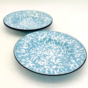 May include: Two round shallow bowls featuring a light blue and white marbled enamel pattern. Each bowl has a smooth, glossy finish and a distinct black rim along the outer edge, creating a clean contrast against the speckled blue and white surface.