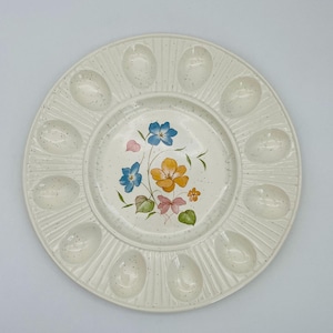 Peut inclure: Assiette à œufs en céramique crème avec un motif floral. L'assiette comporte 12 empreintes en forme d'œuf sur le bord et un motif floral central avec des fleurs bleues, jaunes et roses. La plaque a une finition mouchetée.