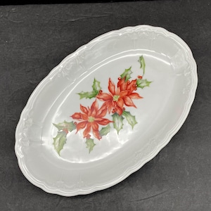 May include: An oval white ceramic serving platter featuring a central decorative illustration of two red poinsettia flowers with green leaves and small red berries. The rim of the platter has a subtle embossed scalloped edge design.