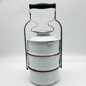 May include: A three-tiered white enamel food carrier with red rim accents. The cylindrical container features a metal frame with a black handle, designed to stack three separate compartments vertically under a single lid with a secure locking mechanism.