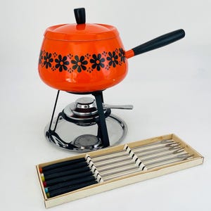 May include: An orange fondue pot with a black floral design, black handle, and lid. The pot sits on a black metal stand with a silver warming plate. A box of fondue forks with black handles and colored tips is also present.