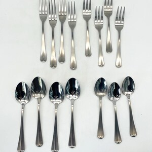 May include: A collection of stainless steel silverware, including forks and spoons, arranged on a white surface. The forks have four tines and the spoons have oval bowls. The silverware has a simple, elegant design.