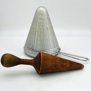 May include: A conical metal food mill strainer with a fine perforated surface and a silver finish, accompanied by a separate wooden pestle featuring a rounded handle and a tapered point, designed for pressing ingredients through the mesh sieve.