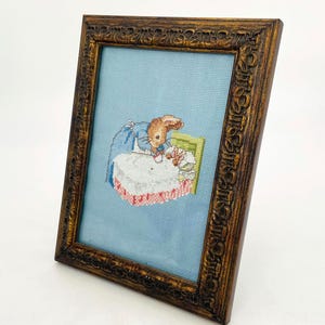 May include: A framed cross-stitch depicting a rabbit in a blue jacket and a white apron, seated at a table. The artwork is set against a light blue background and framed in a dark brown, ornate wooden frame. The frame measures approximately 8 x 10 inches.