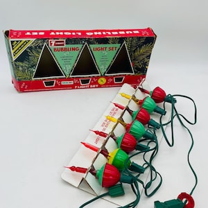 Può includere: Un set di luci natalizie vintage con sette luci a bolle multicolori. Le luci sono rosse, verdi e gialle, con un cavo e una spina verdi. La scatola è rossa e riporta la scritta "Bubbling Light Set".