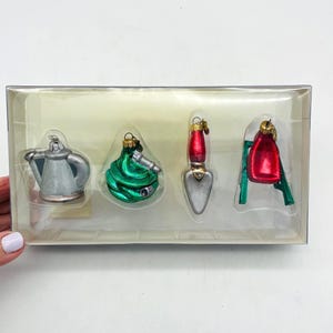 May include: A set of four miniature garden-themed ornaments in a clear plastic package. The ornaments include a silver watering can, a green hose, a trowel with a red handle, and a red watering can on a green ladder.