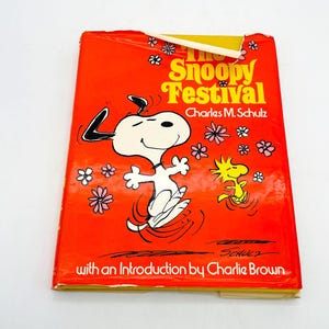 May include: A vintage book titled "The Snoopy Festival" by Charles M. Schulz. The cover is bright orange with illustrations of Snoopy, Woodstock, and flowers. The book includes an introduction by Charlie Brown.