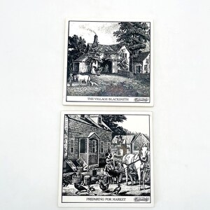 May include: Two black and white square coasters with detailed illustrations of village scenes. One coaster depicts "THE VILLAGE BLACKSMITH," and the other shows "PREPARING FOR MARKET." The coasters feature buildings, animals, and people.
