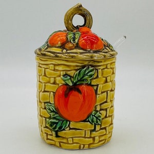 May include: A ceramic jar with a lid, featuring a basket weave design in yellow and brown. The jar is adorned with a large red apple and green leaves. The lid is topped with orange fruit and a loop handle. A clear spoon rests on the side.
