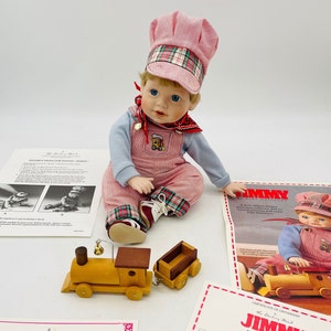 Peut inclure: Une poupée en porcelaine nommée "Jimmy" vêtue d'une tenue en velours côtelé rose, d'un chapeau assorti avec des carreaux et des chaussures blanches. Un train jouet en bois et des papiers sont également présents. La poupée a les yeux bleus et les cheveux blonds.